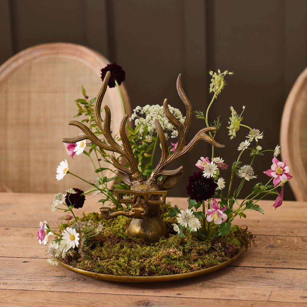 A decorative brass deer head arrangement on a round tray, surrounded by green moss and small wildflowers, set on a wooden table with woven-back chairs in the background.
