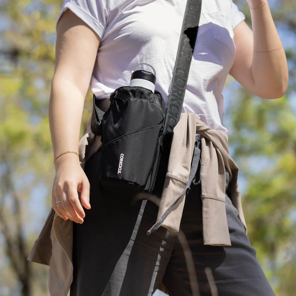 Person wearing a black waist bag with a visible brand logo, standing outdoors with trees in the background.