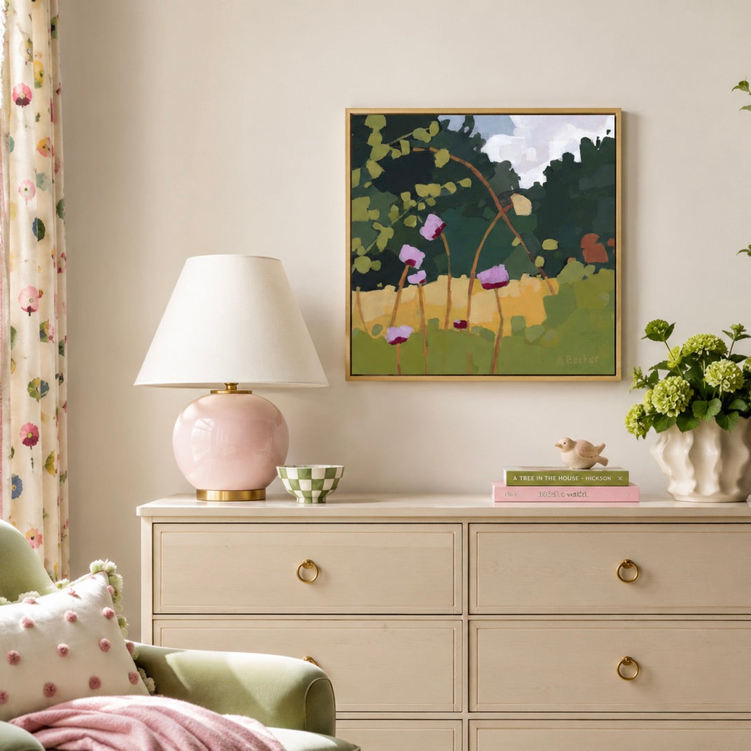 A framed abstract landscape painting with green foliage, purple flowers, and a blue sky, displayed on a cream wall above a light wood dresser in a styled room setting.