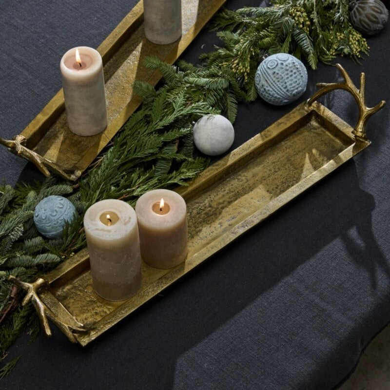 Two long rectangular brass trays with antler handles, styled with green pine branches, textured blue and white decorative balls, and two lit cream pillar candles on a dark tablecloth.