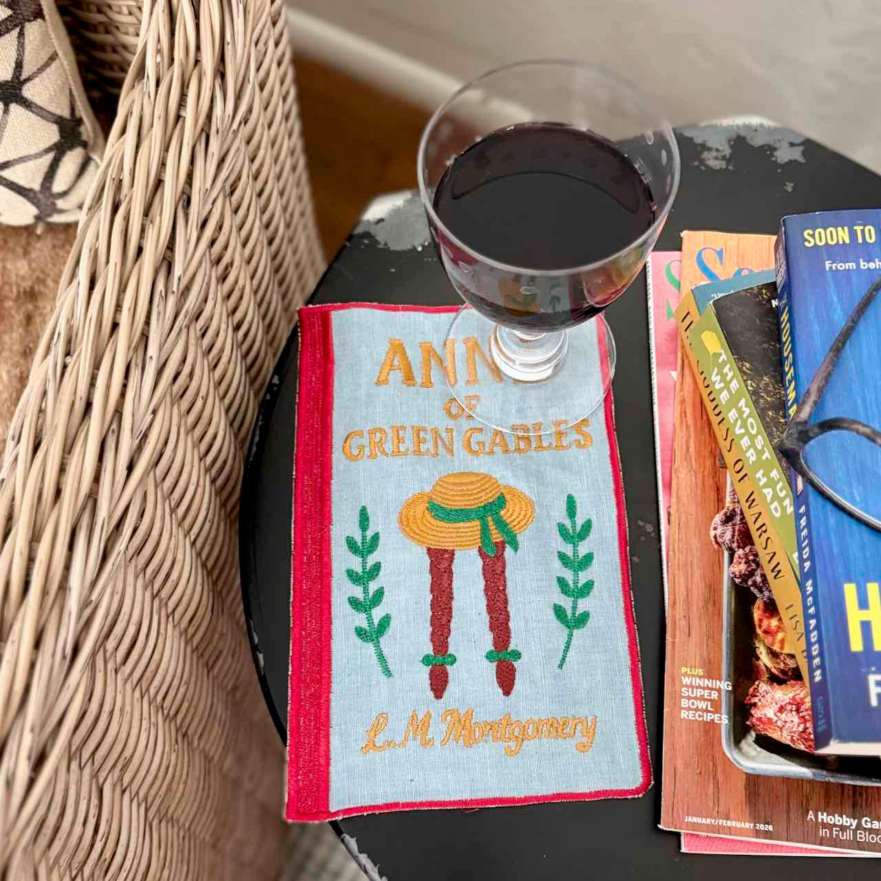 Embroidered towel with 'Anne of Green Gables' design on a table with a glass of red wine and books.