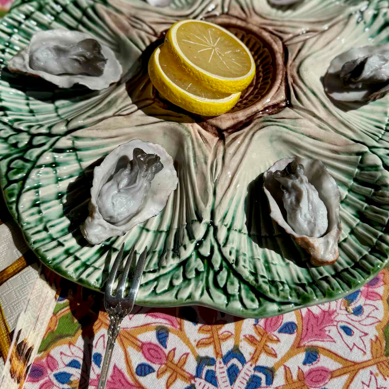 A green and beige ceramic oyster shell platter with a leaf-like embossed pattern, served with four raw oysters and two lemon slices on a patterned tablecloth.