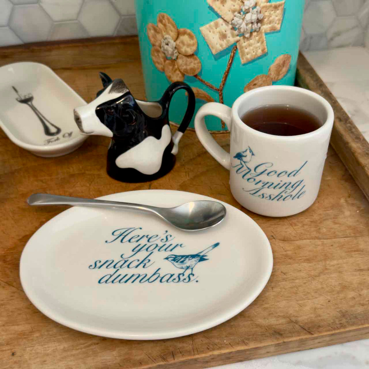 A styled tabletop scene with a white ceramic plate printed with the phrase 'Here's your snack dumbass.' and a matching white mug printed with 'Good Forgetting Brisket.