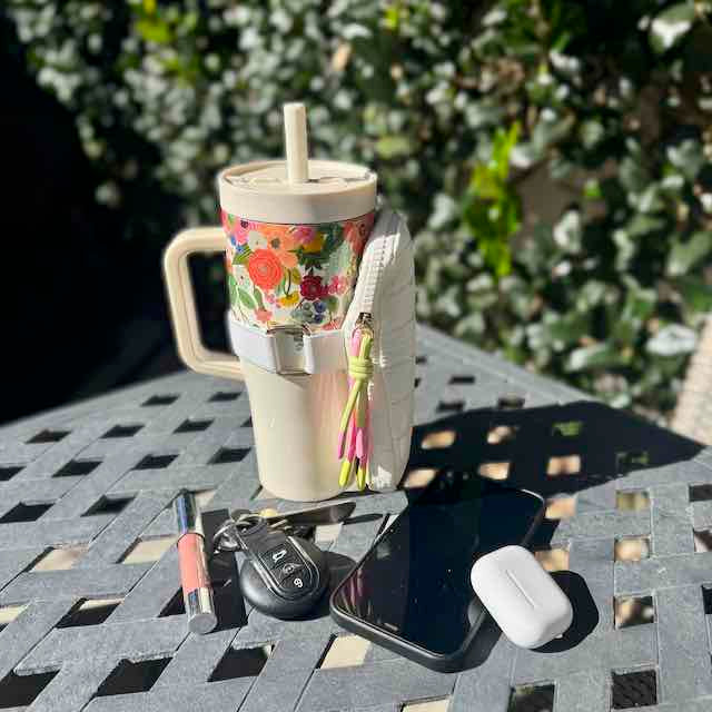 Floral-patterned tumbler with a straw, phone, keys, and lip gloss on a table outdoors.