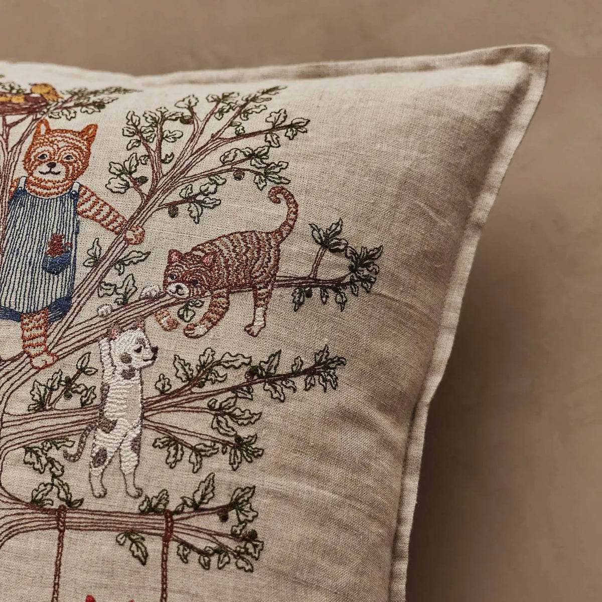 A close-up of a beige linen throw pillow featuring embroidered cats and a dog climbing a tree, set against a matching beige background.