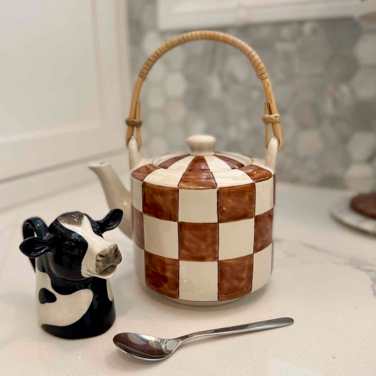 Checkered teapot with a cow-shaped salt shaker and spoon on a marble surface.