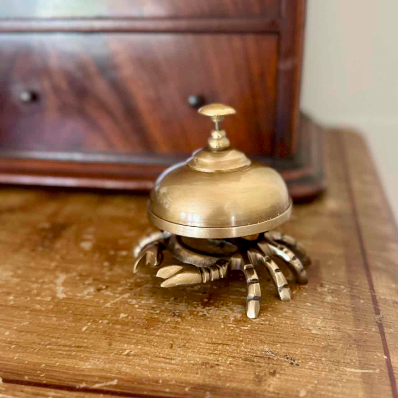 A brass crab-shaped bell holder with a dome-shaped top and metallic claws, resting on a worn wooden surface with a blurred wooden cabinet in the background.