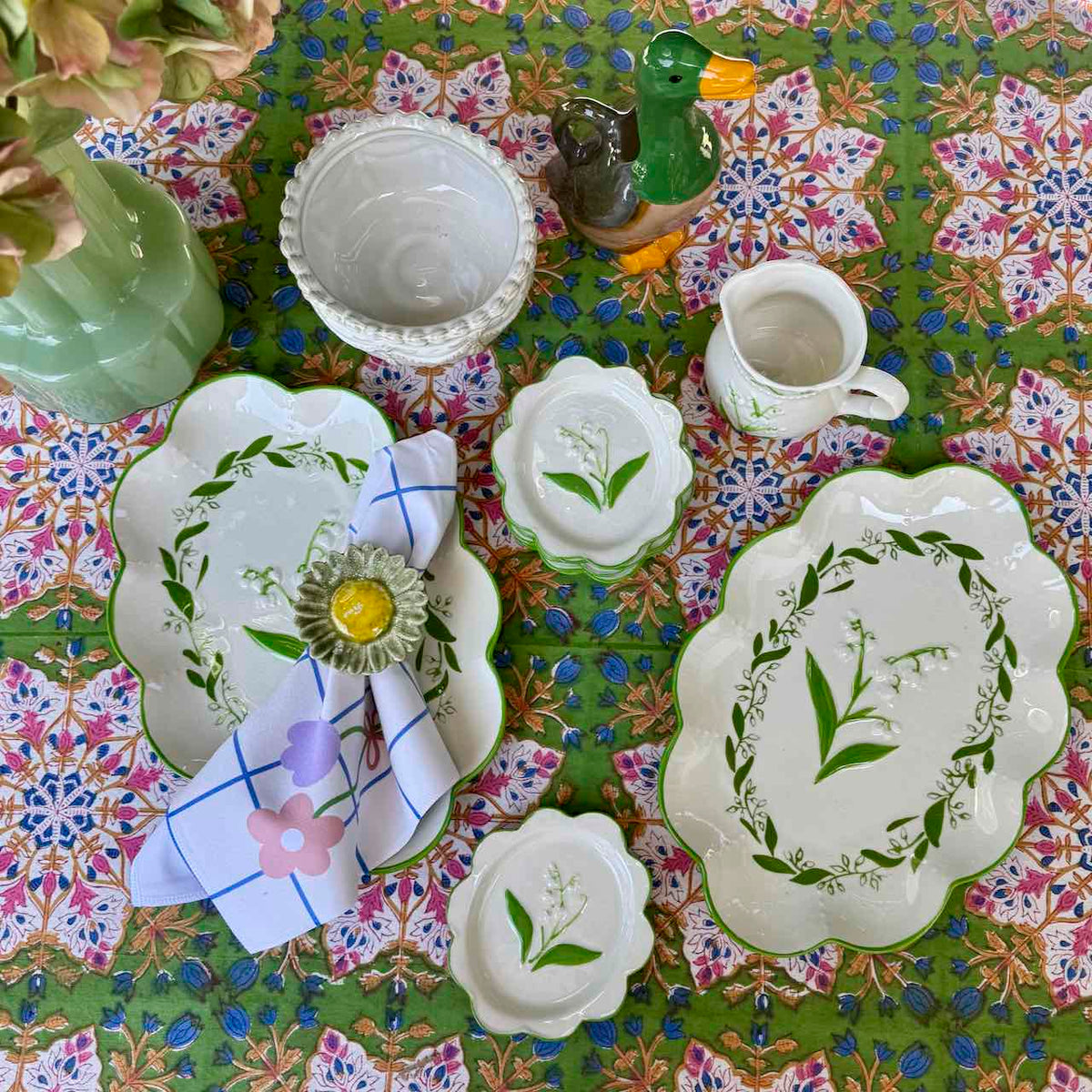 A coordinated set of white ceramic tableware with green botanical illustrations, arranged on a colorful floral tablecloth. The set includes platters, bowls, a small pitcher, and a napkin with a flower centerpiece.