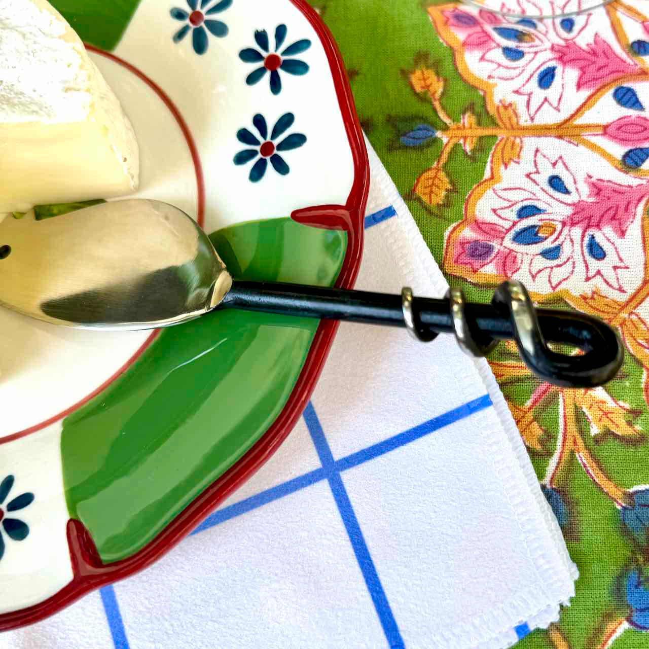 A close-up of a decorative ceramic plate with a green leaf-shaped center, blue floral motifs, and a red rim, holding a piece of cheese and a spoon with a black handle. A white napkin with blue grid lines and a green patterned fabric are visible underneath.