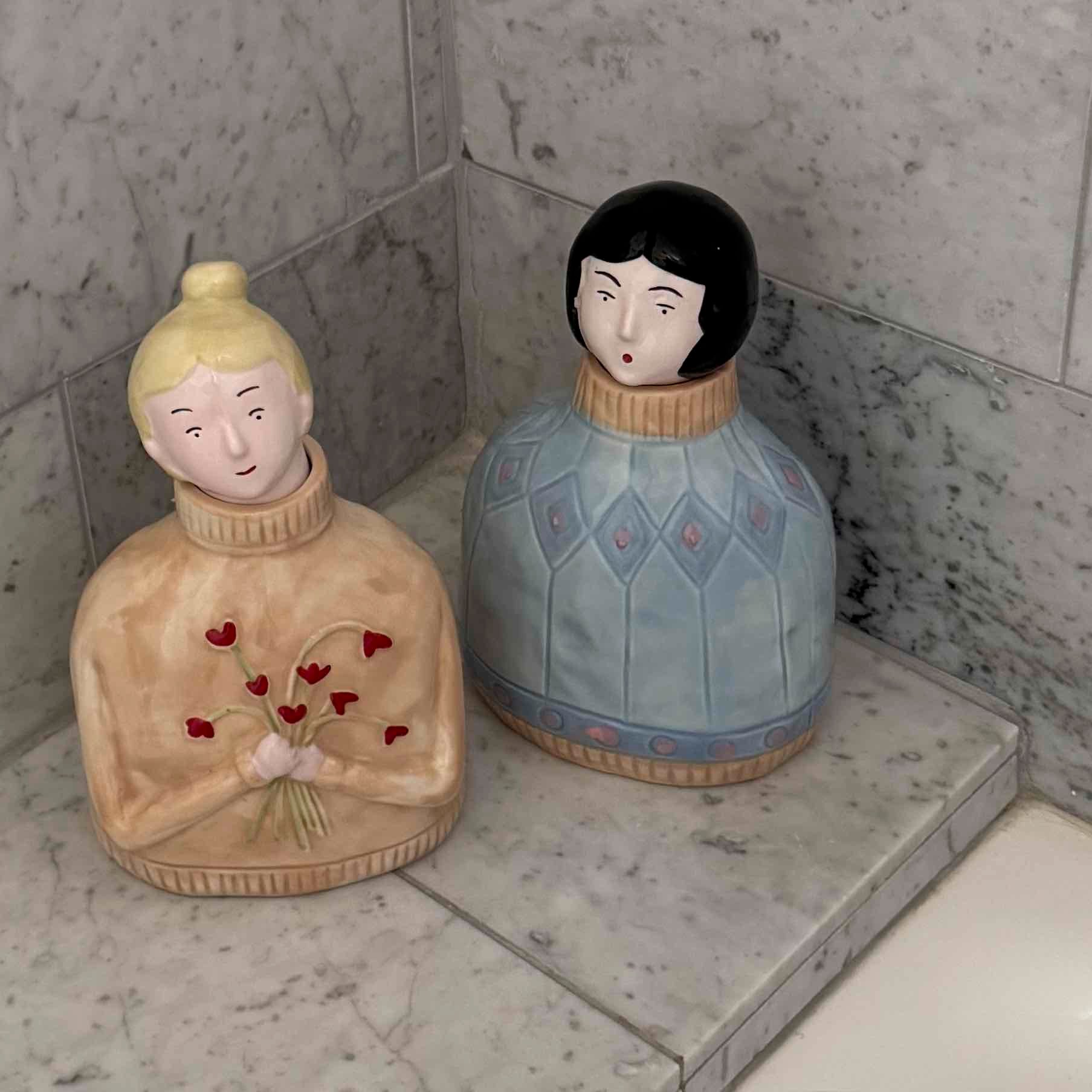 Two ceramic figurines on a marble surface with a tiled wall background