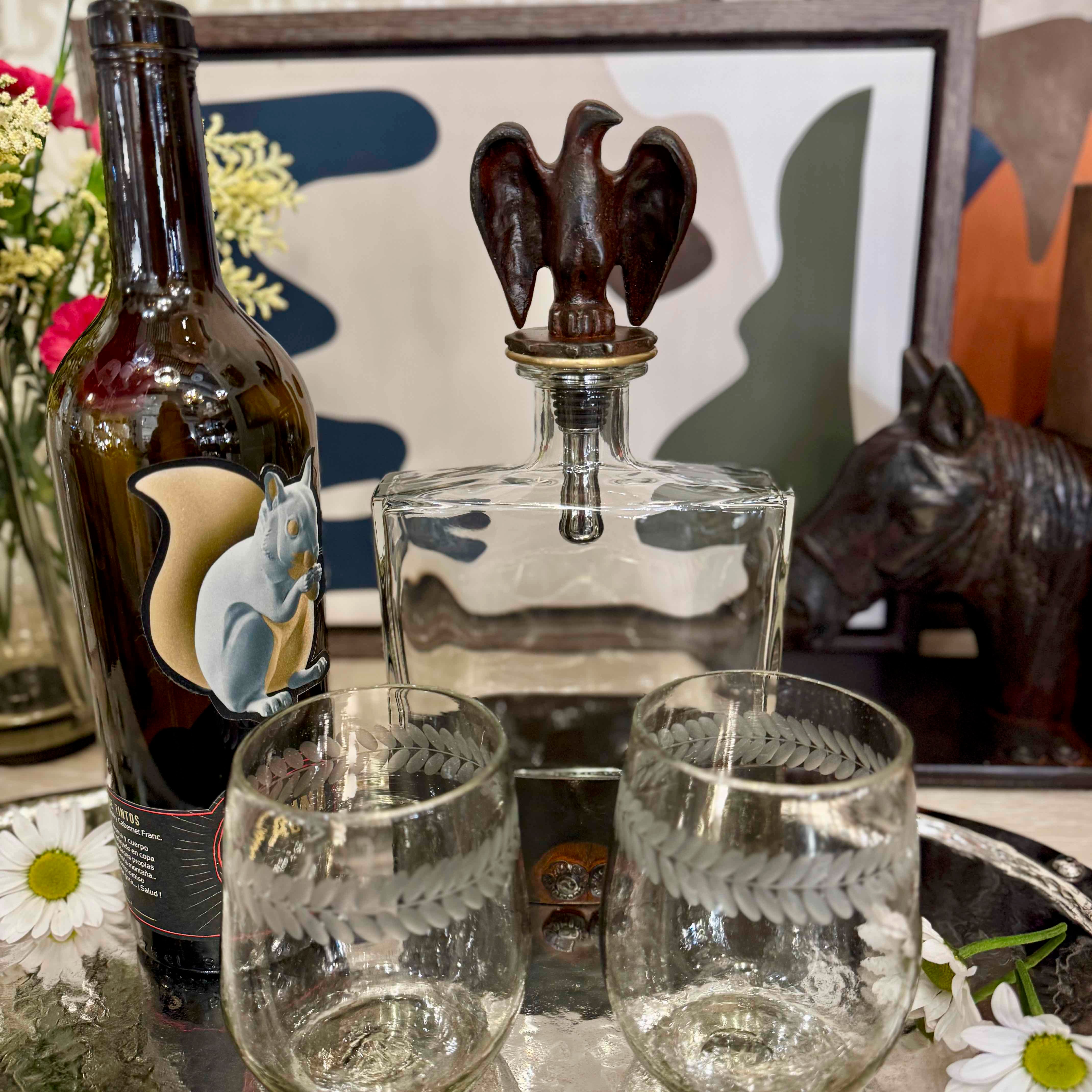 Decanter with wooden stopper, bottle, and glasses on a decorative surface with flowers and artwork.