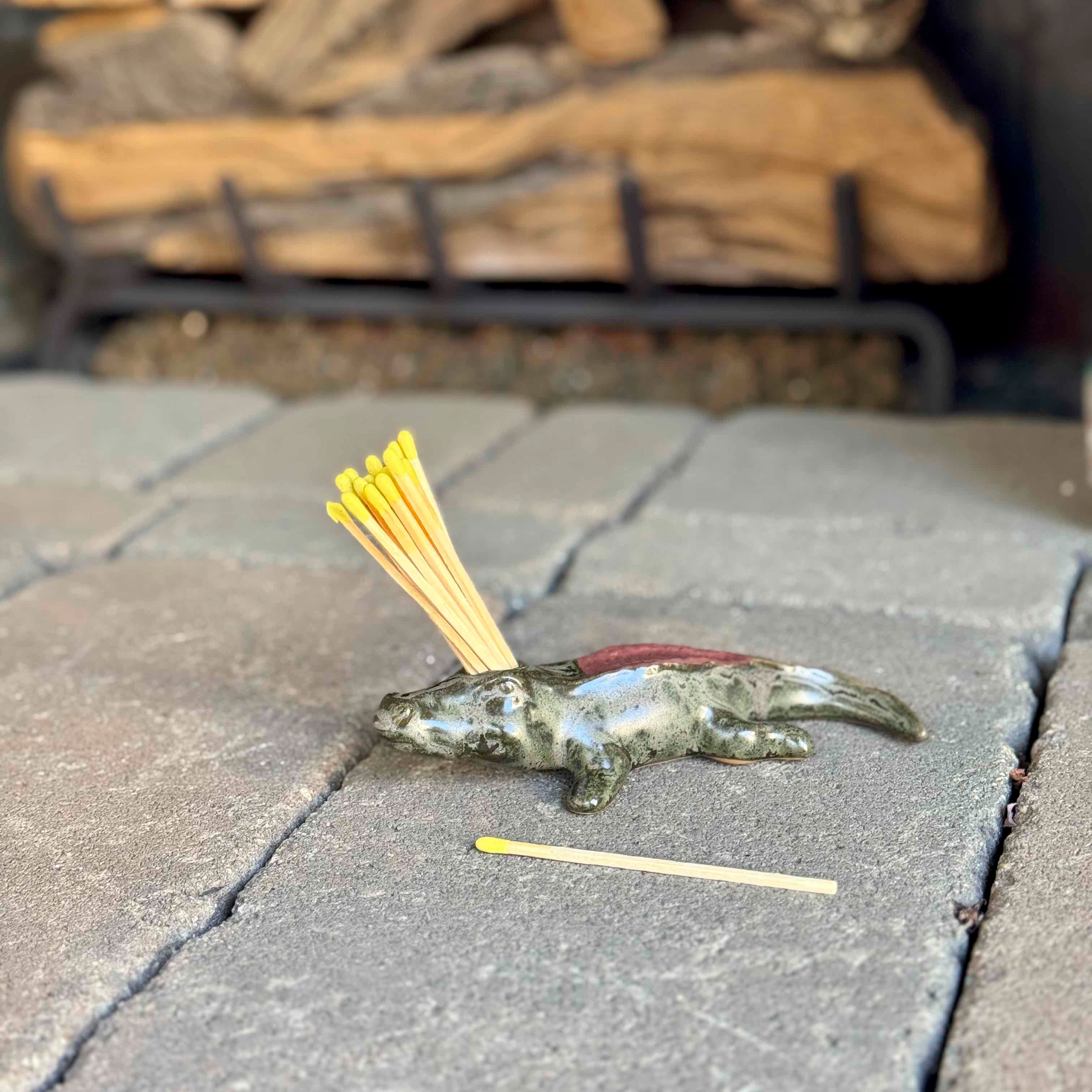 Small lizard figurine with yellow sticks on a stone surface, with stacked firewood in the background.