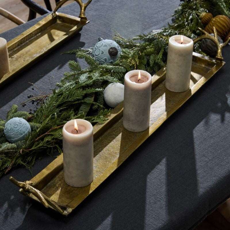 Three lit pillar candles arranged on a long gold tray with evergreen branches and decorative ornaments on a dark tablecloth.