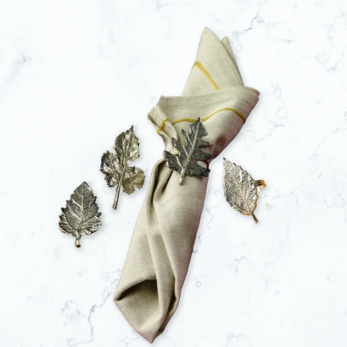 A beige linen napkin with gold stripes and a metallic leaf-shaped napkin ring, arranged on a white marble surface alongside three matching leaf-shaped metal accents.