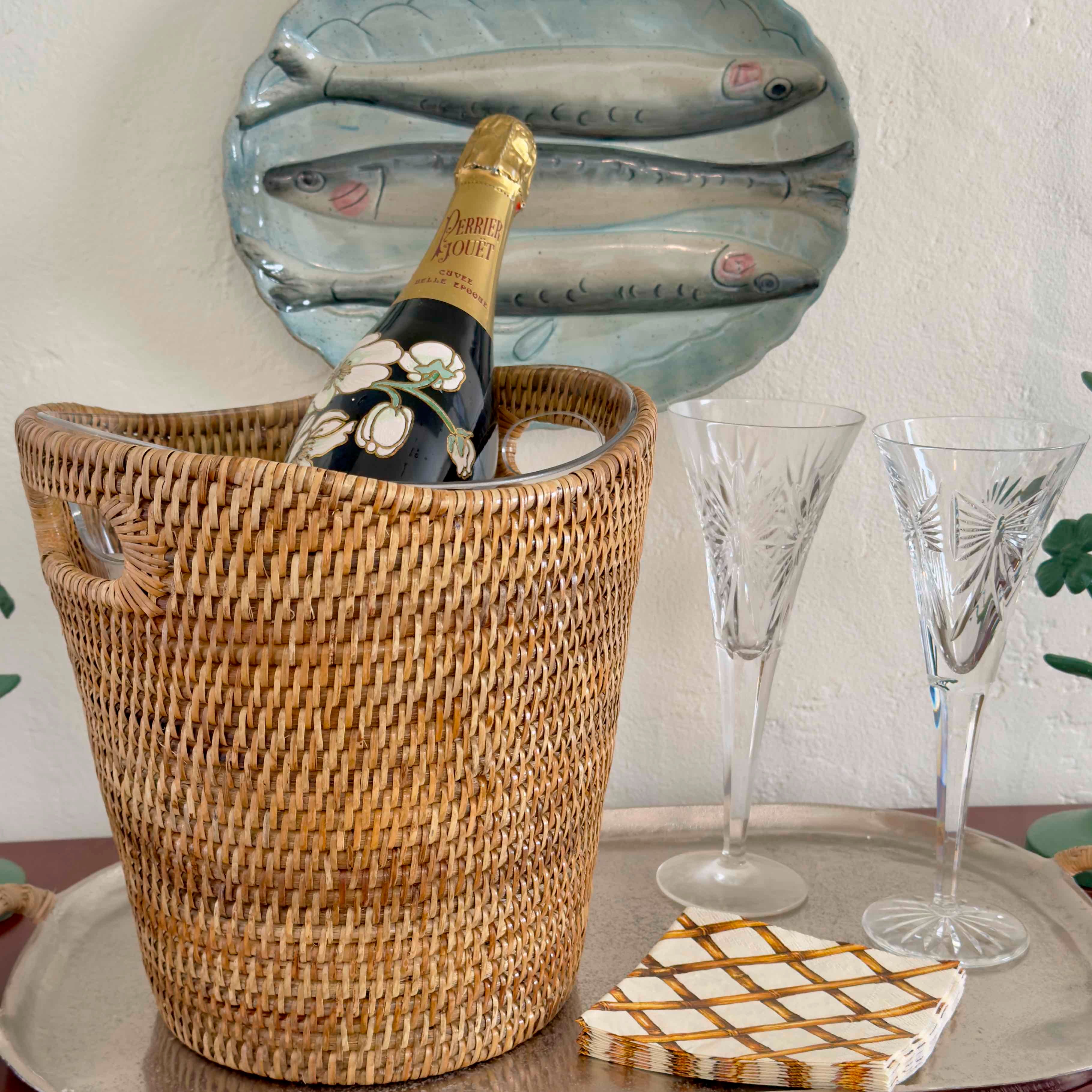 Wicker wine cooler with bottle of champagne, two glasses, and a decorative plate on a table.