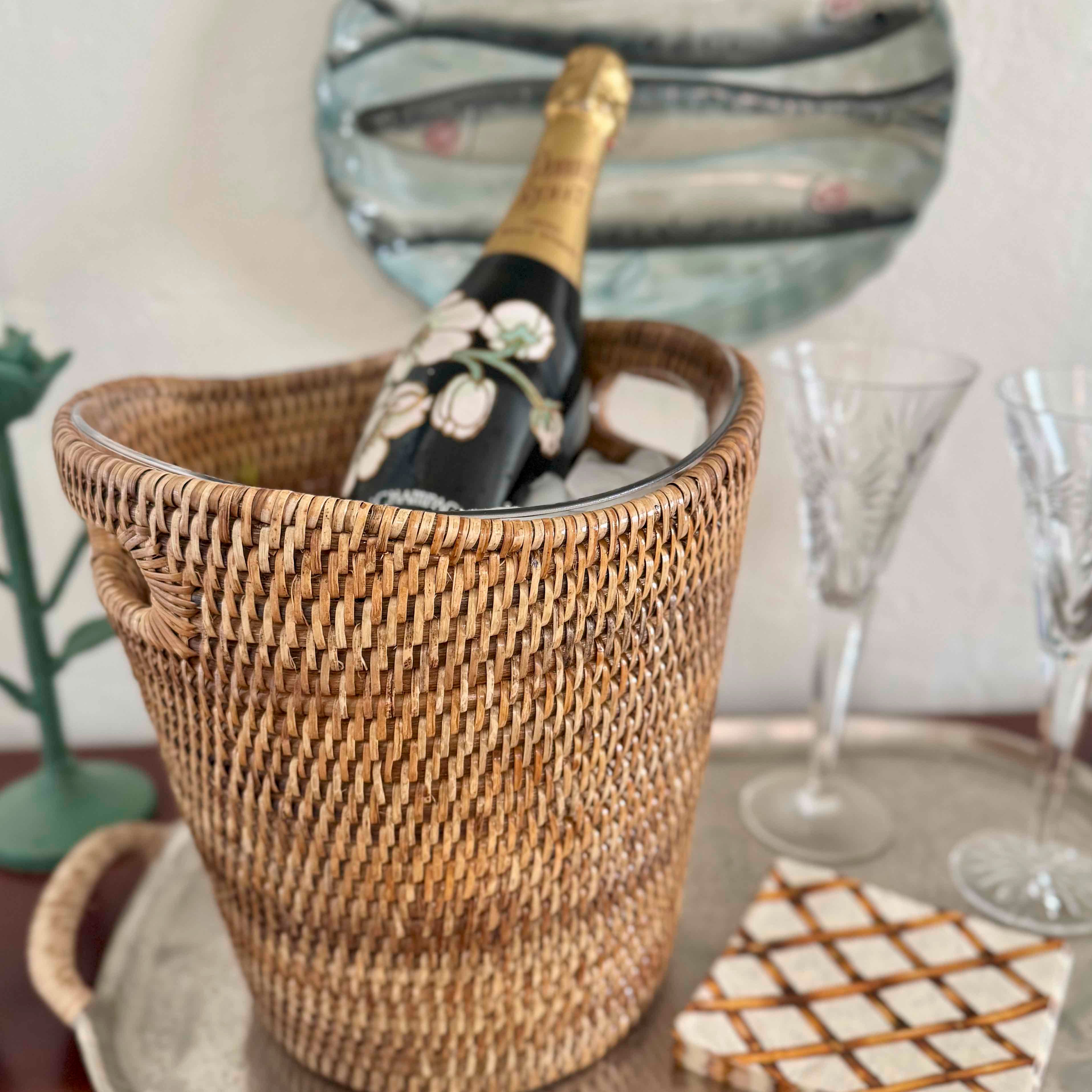 Wicker wine cooler with a bottle of champagne on a table with glasses and a mirror in the background.