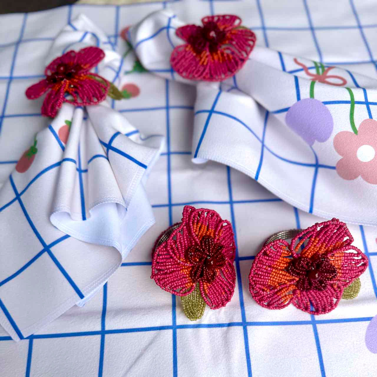 Three beaded flower-shaped napkin rings in pink, red, and orange with green leaves, arranged on a white tablecloth with a blue grid pattern and floral prints.