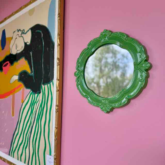 Green decorative mirror on a pink wall with a painting to the left.