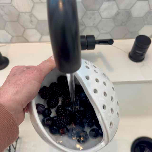 Person washing blackberries in a white colander under running water.