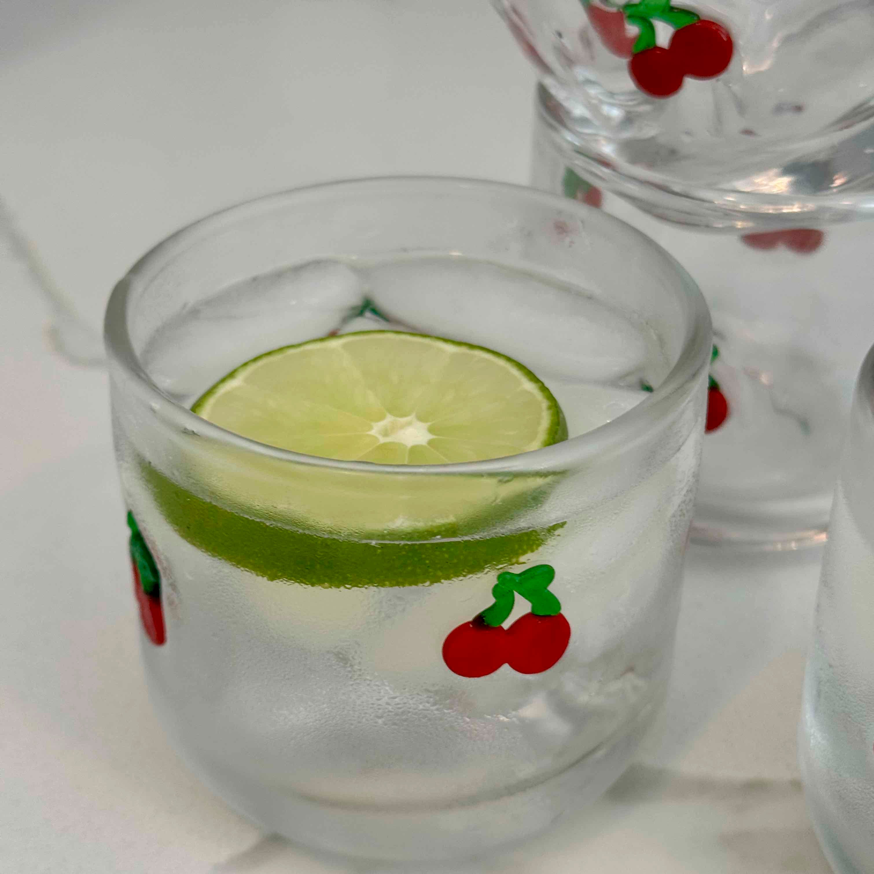 Clear glass with ice and lime slice, decorated with cherries on a white surface