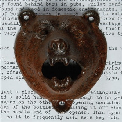 A rustic cast iron bear head bottle opener with a weathered, rusted brown finish, displayed against a background of printed text.