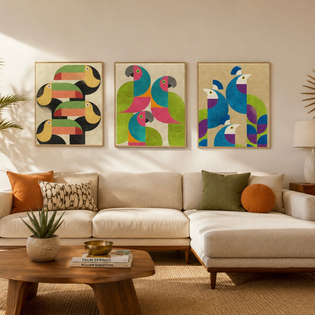 Three framed abstract bird wall art prints hang above a beige sectional sofa in a styled living room. The artwork features colorful toucans, parrots, and peacocks in green, blue, pink, yellow, and black on cream backgrounds.