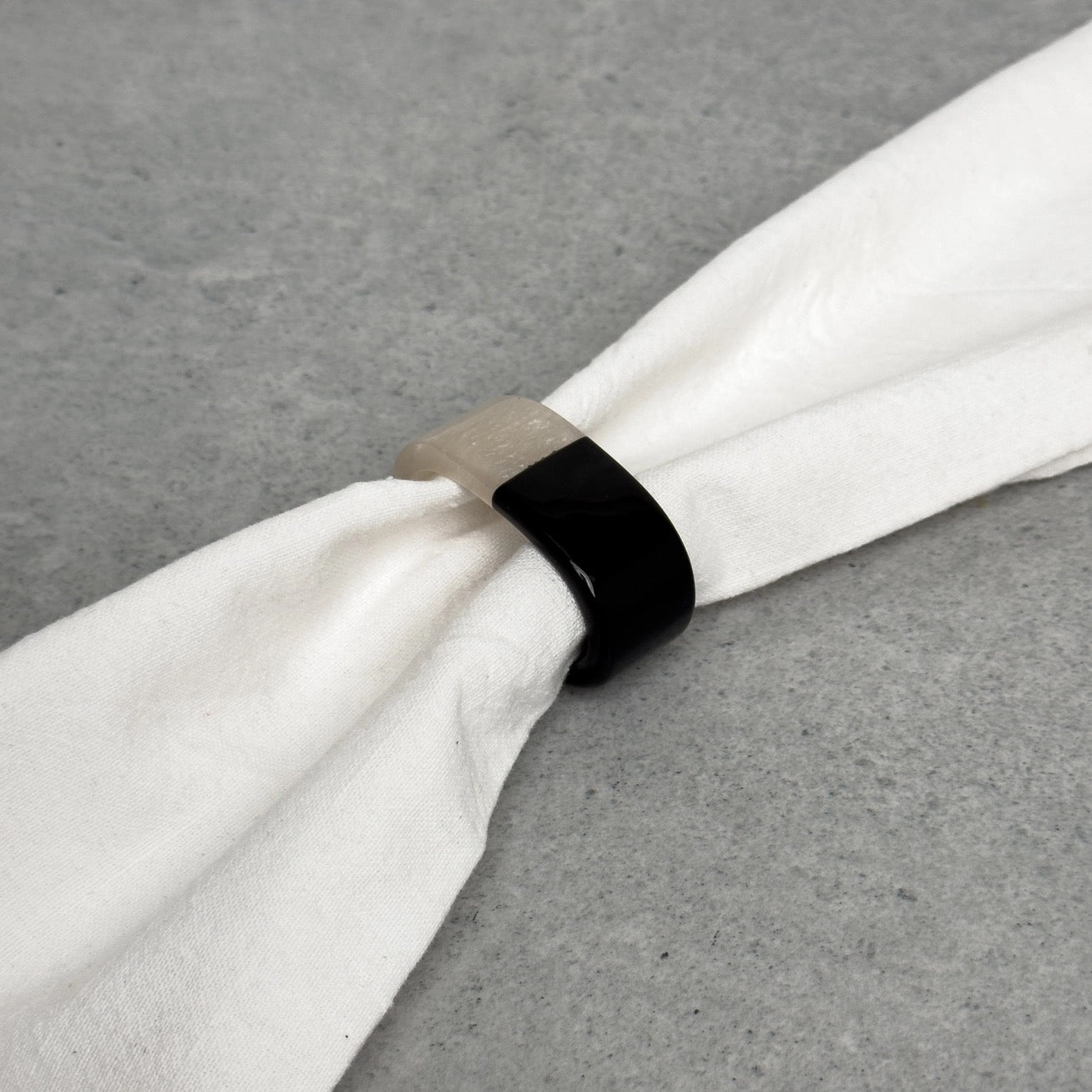 A black ring-shaped napkin holder with a light beige textured end, shown holding a white napkin on a gray textured surface.