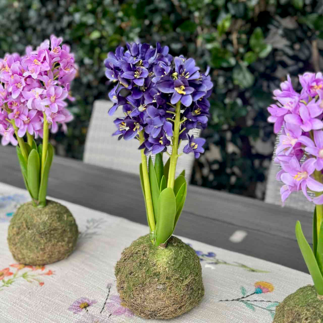Three small artificial wisteria flower arrangements in moss balls on a table outdoors, with greenery and patio chairs blurred in the background.