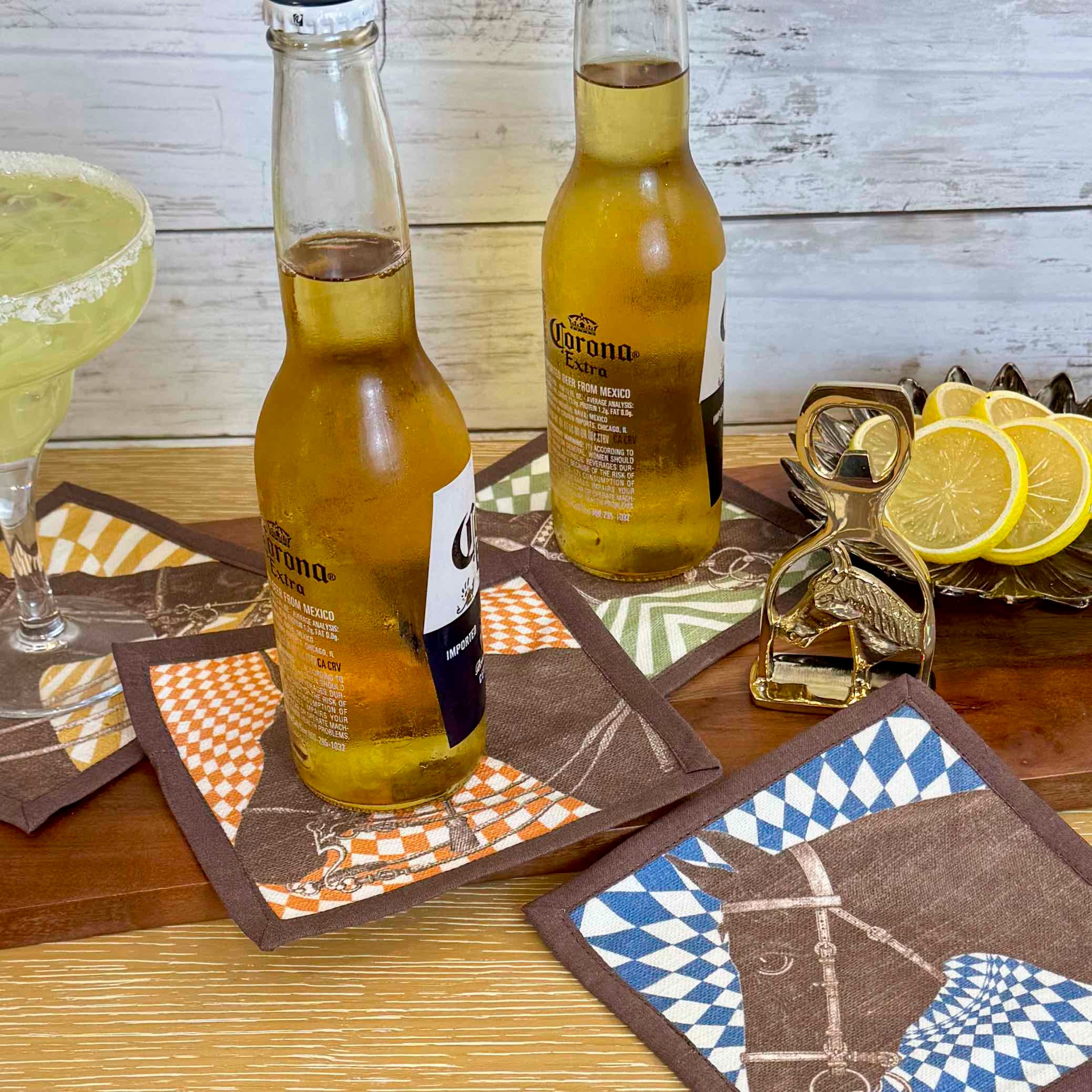 Two bottles of Corona beer on patterned coasters with a glass of green drink and lemon slices in the background.