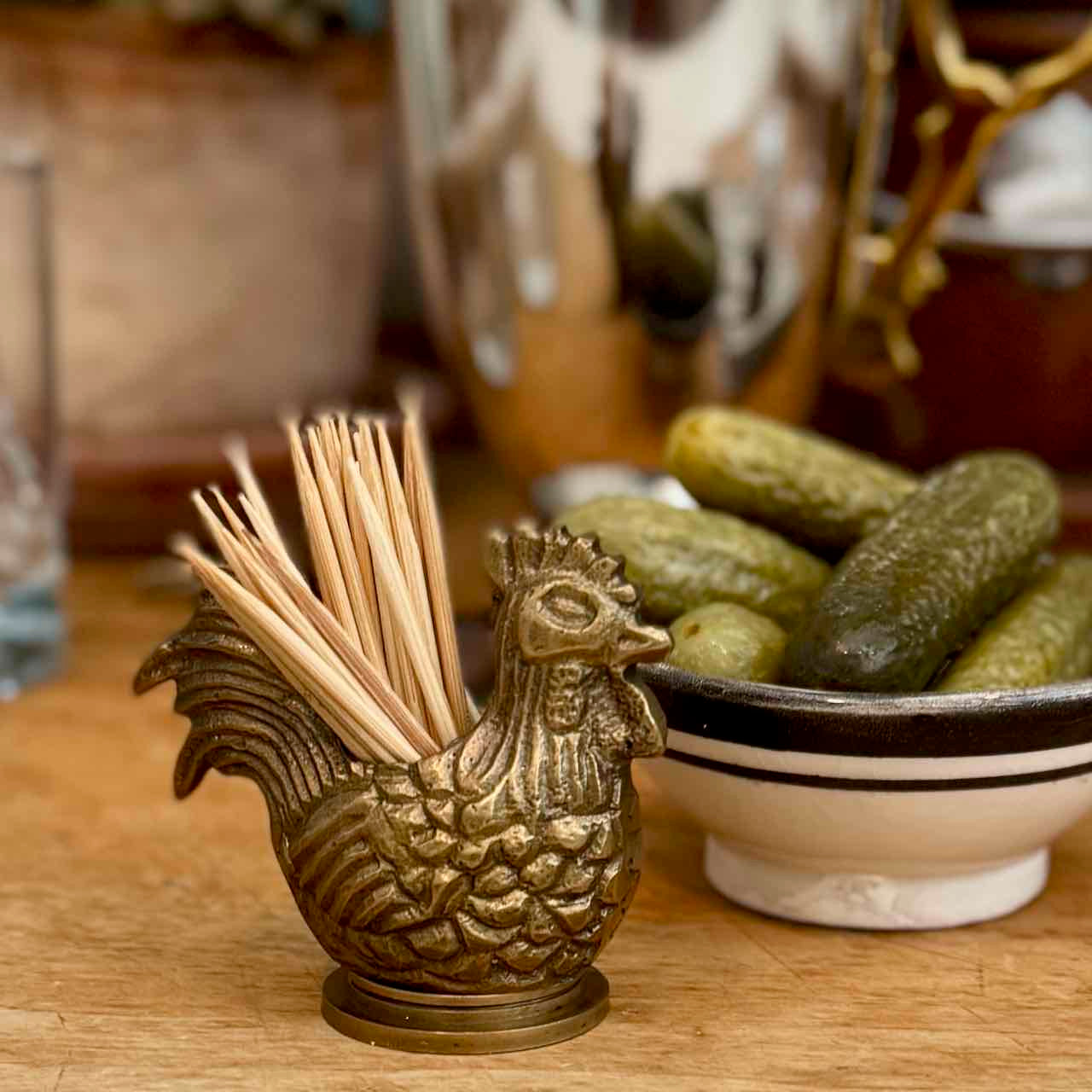 A brass rooster-shaped toothpick holder filled with wooden toothpicks, set on a wooden table next to a bowl of pickles in a warm, softly blurred kitchen setting.