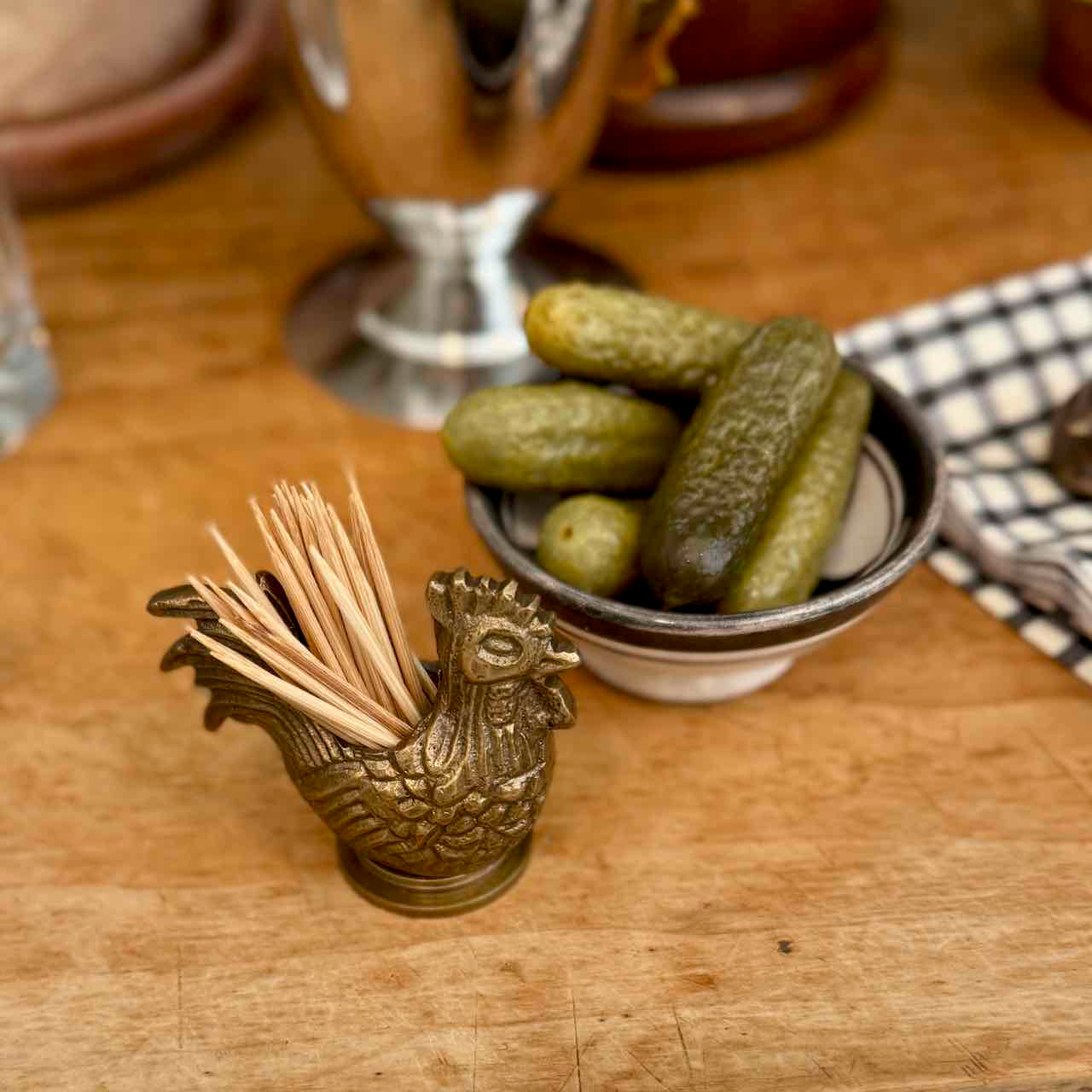 A brass rooster-shaped toothpick holder filled with wooden toothpicks sits on a wooden tabletop next to a small bowl of pickles.
