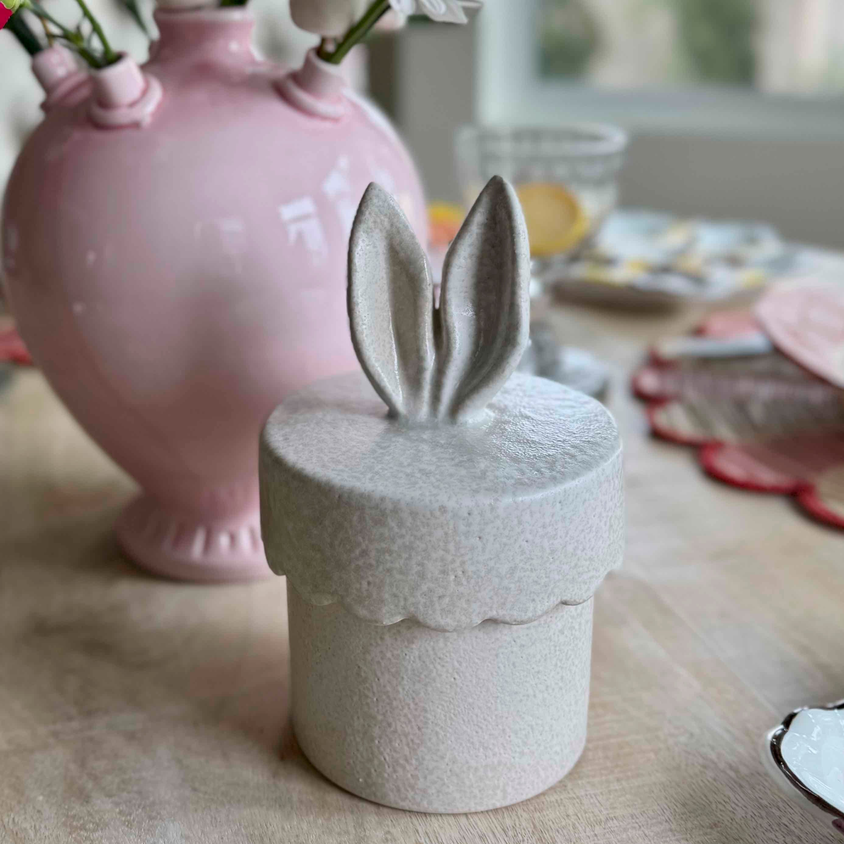 Small white decorative item with bunny ears on a table next to a pink vase.