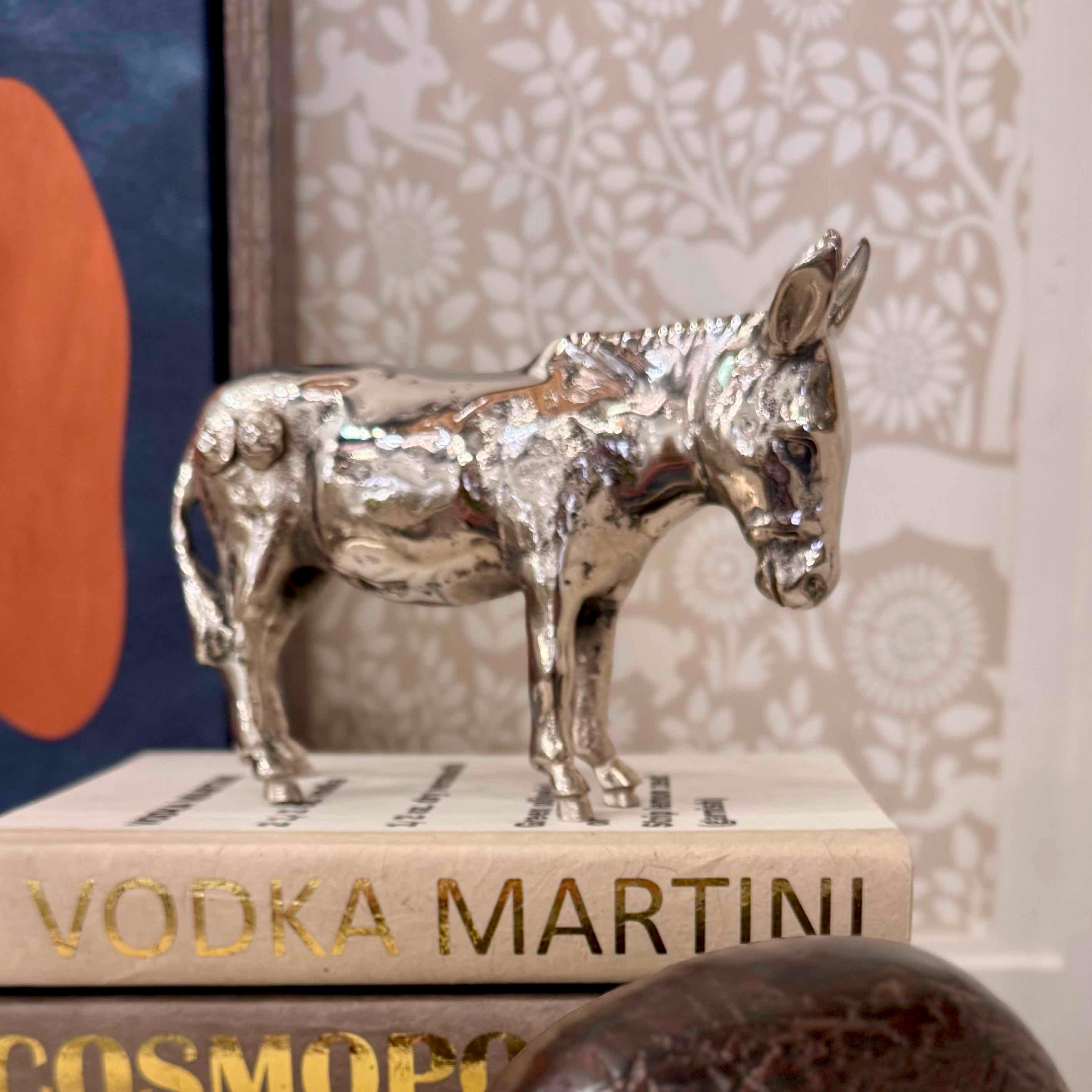 Silver donkey figurine on top of a book titled 'Vodka Martini' with a decorative wall in the background.