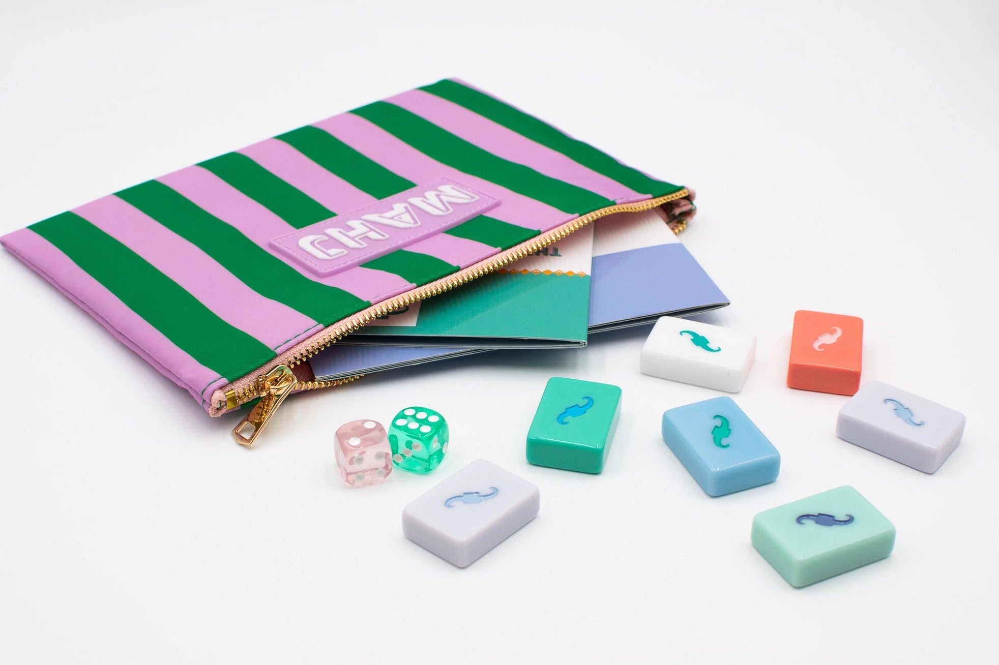 A striped zip pouch in pink and green with a gold zipper, shown open with several pastel game pieces and dice scattered in front on a white background.