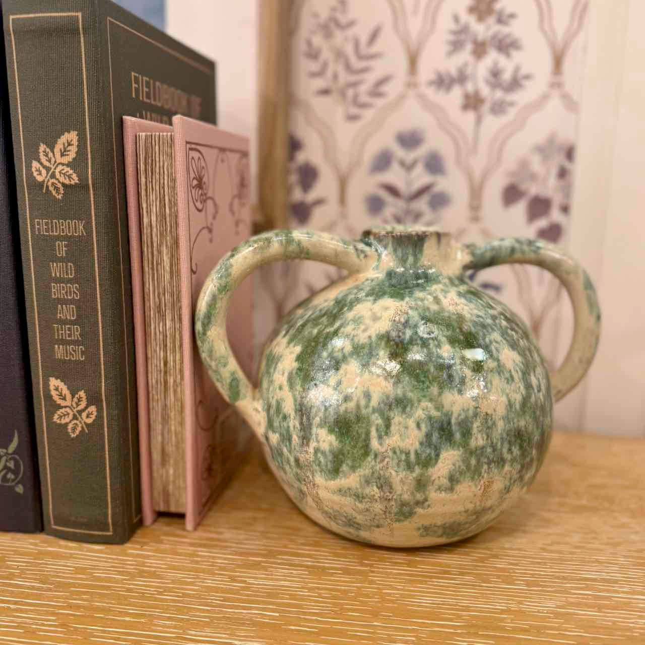 Green ceramic vase with two handles on a wooden surface next to books.