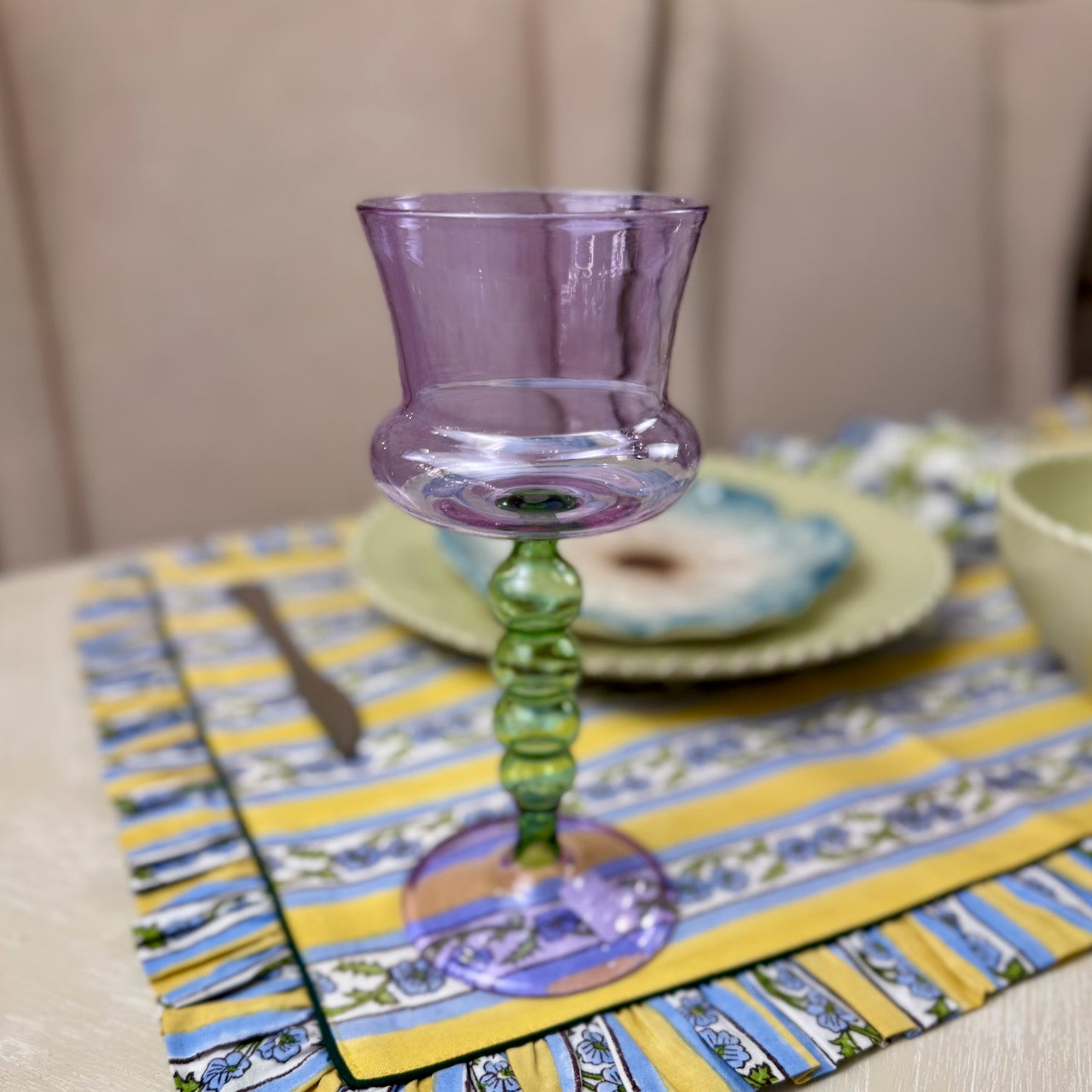 Colorful glass candle holder on a patterned tablecloth with plates and cutlery.