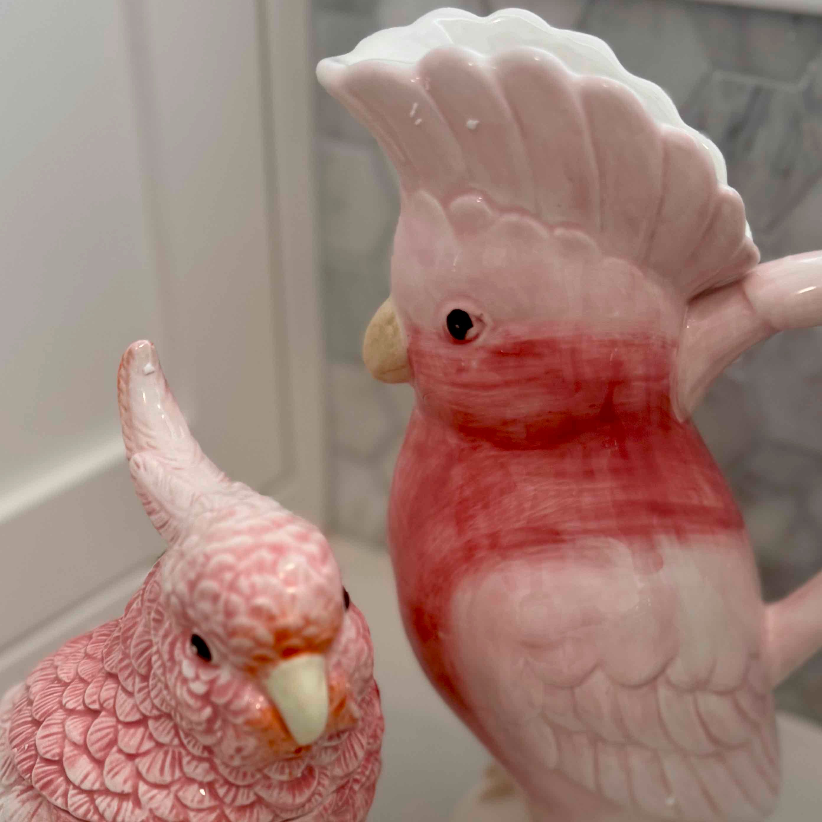 Two ceramic parrot figurines, one larger than the other, on a neutral background.