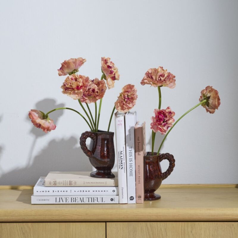 Two small dark brown ceramic pitchers with loop handles, each holding a bouquet of pink and peach carnations, styled on a light wood cabinet with books against a plain light gray wall.