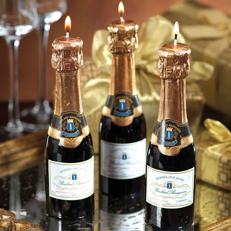 Three miniature sparkling wine bottles with lit candles on top, resembling champagne bottles, on a reflective surface.