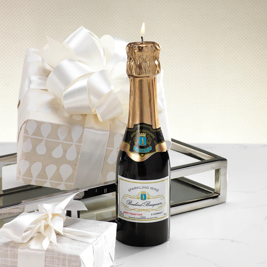 Bottle of sparkling wine with a lit candle on top, next to a wrapped gift with a white bow.