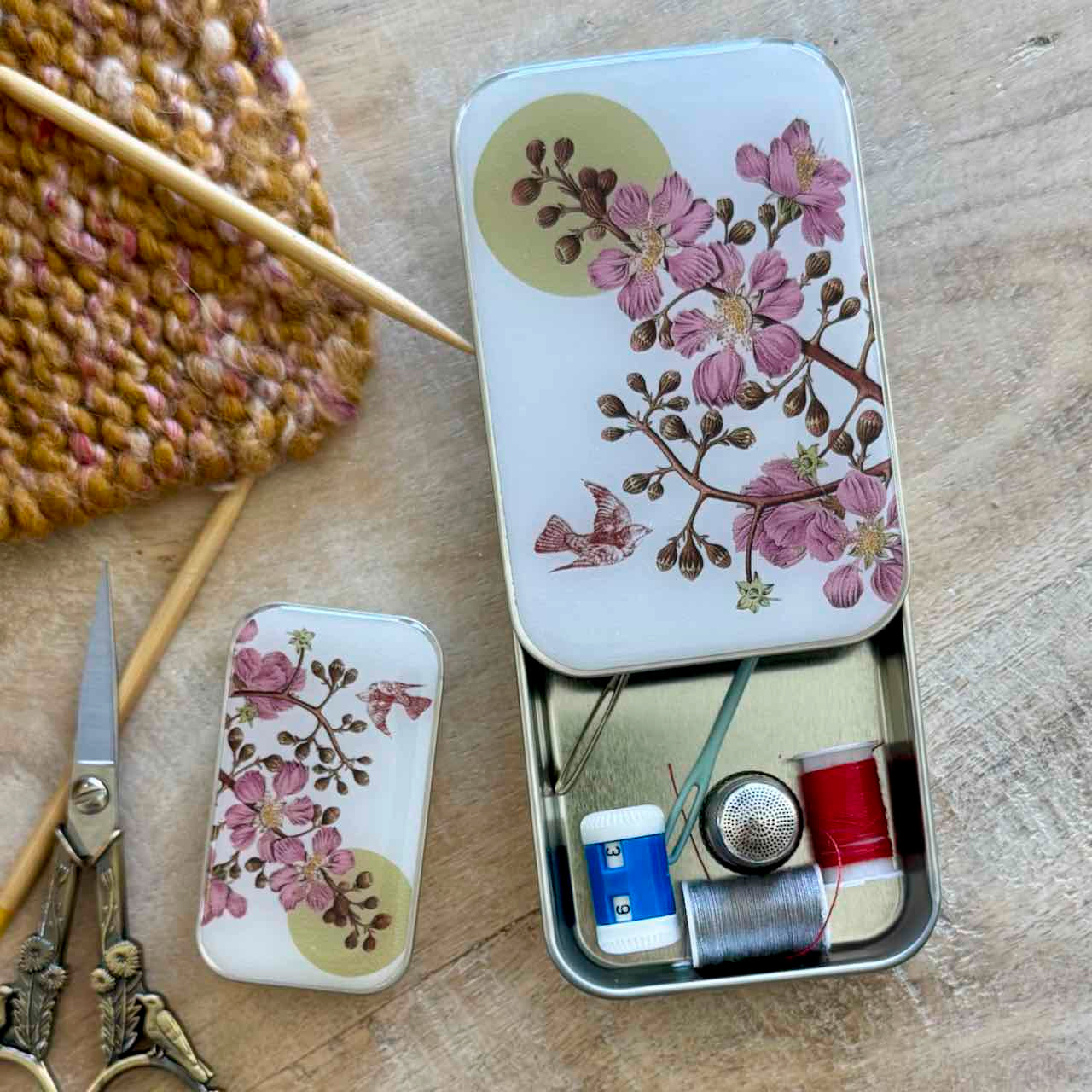 A decorative metal sewing kit tin with a floral print lid, shown open on a wooden surface alongside scissors, knitting needles, and thread spools.