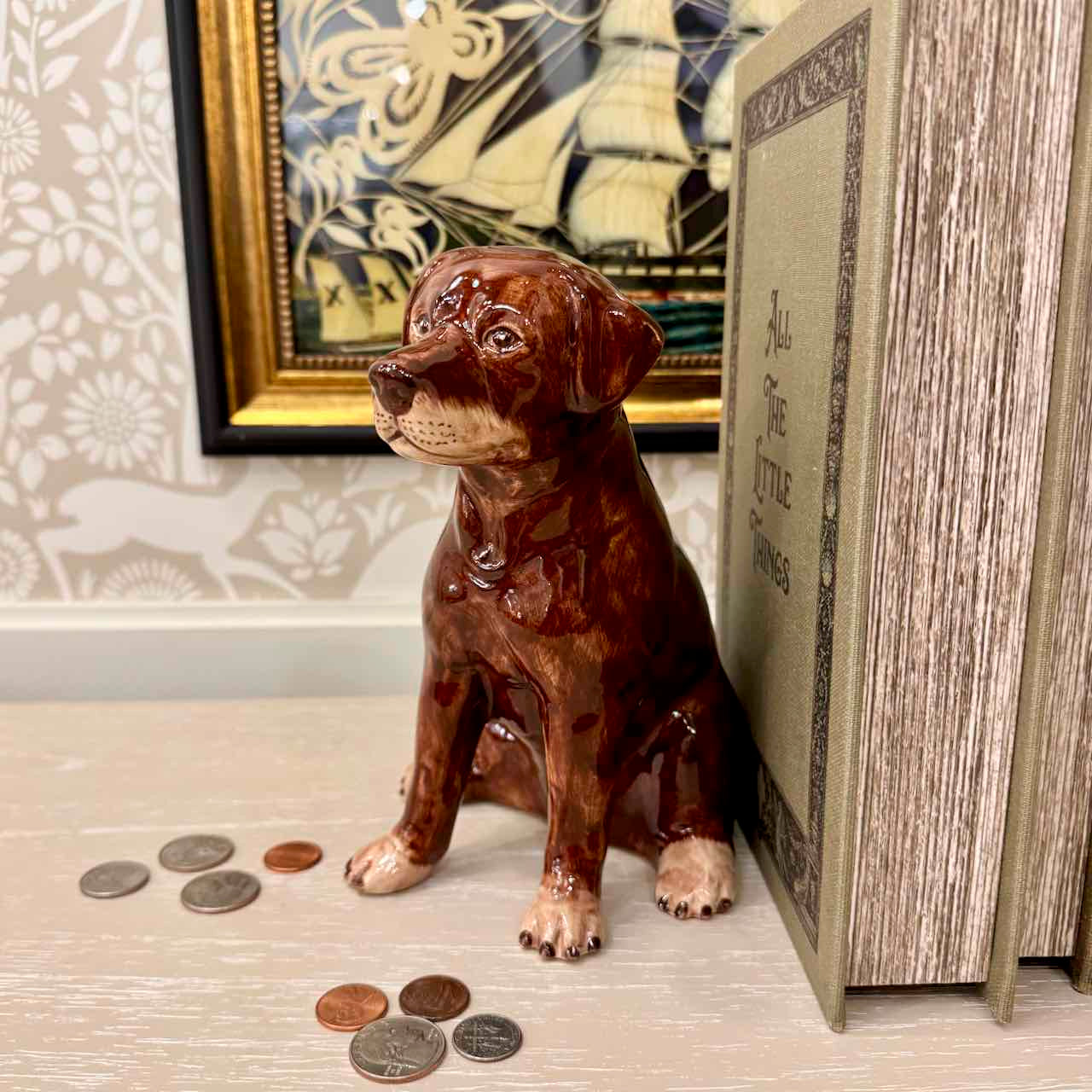 Brown ceramic dog figurine with coins on a surface, next to a book and framed picture.