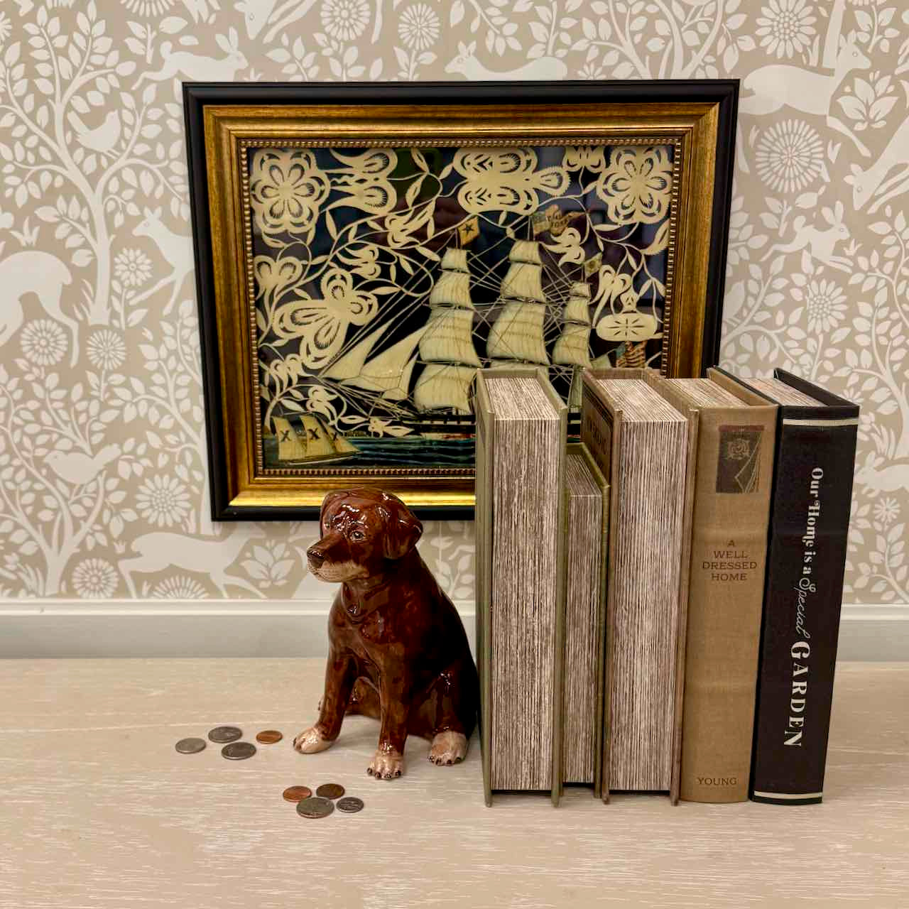 Decorative setting with a framed artwork, books, and a dog figurine on a patterned wall background.