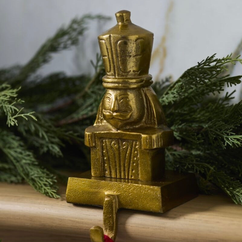 A small gold-toned nutcracker figurine or decorative ornament with a face and crown-like top, displayed on a wooden surface with green evergreen branches in the background.
