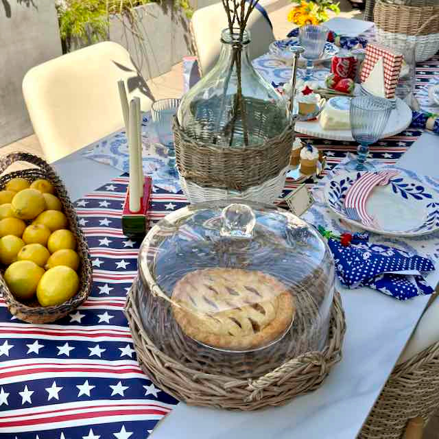 Outdoor table setting with a pie in a wicker basket on a tablecloth with American flag design.