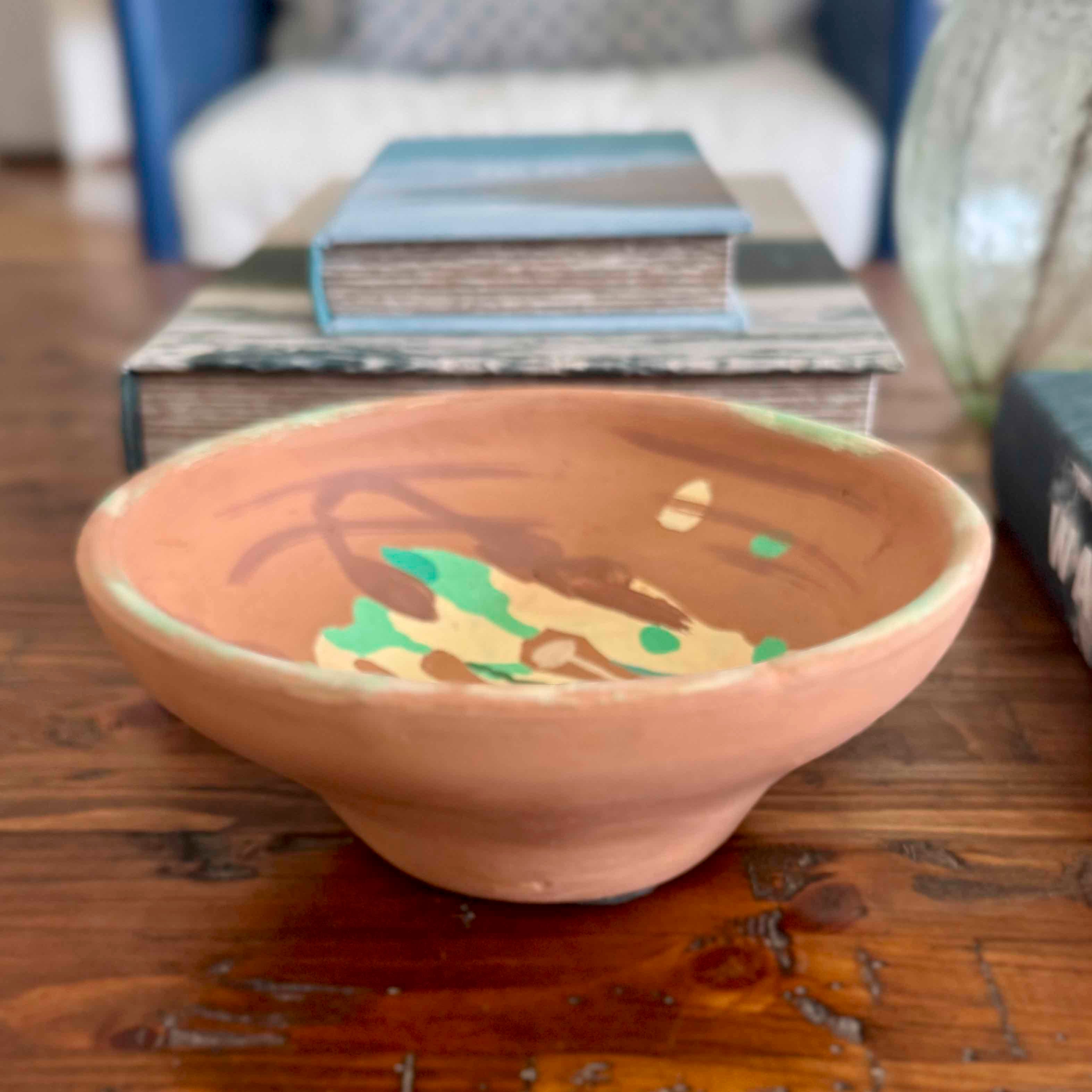A handmade terracotta ceramic bowl with hand-painted abstract designs in brown, green, and beige, resting on a rustic wooden table with books in the background.