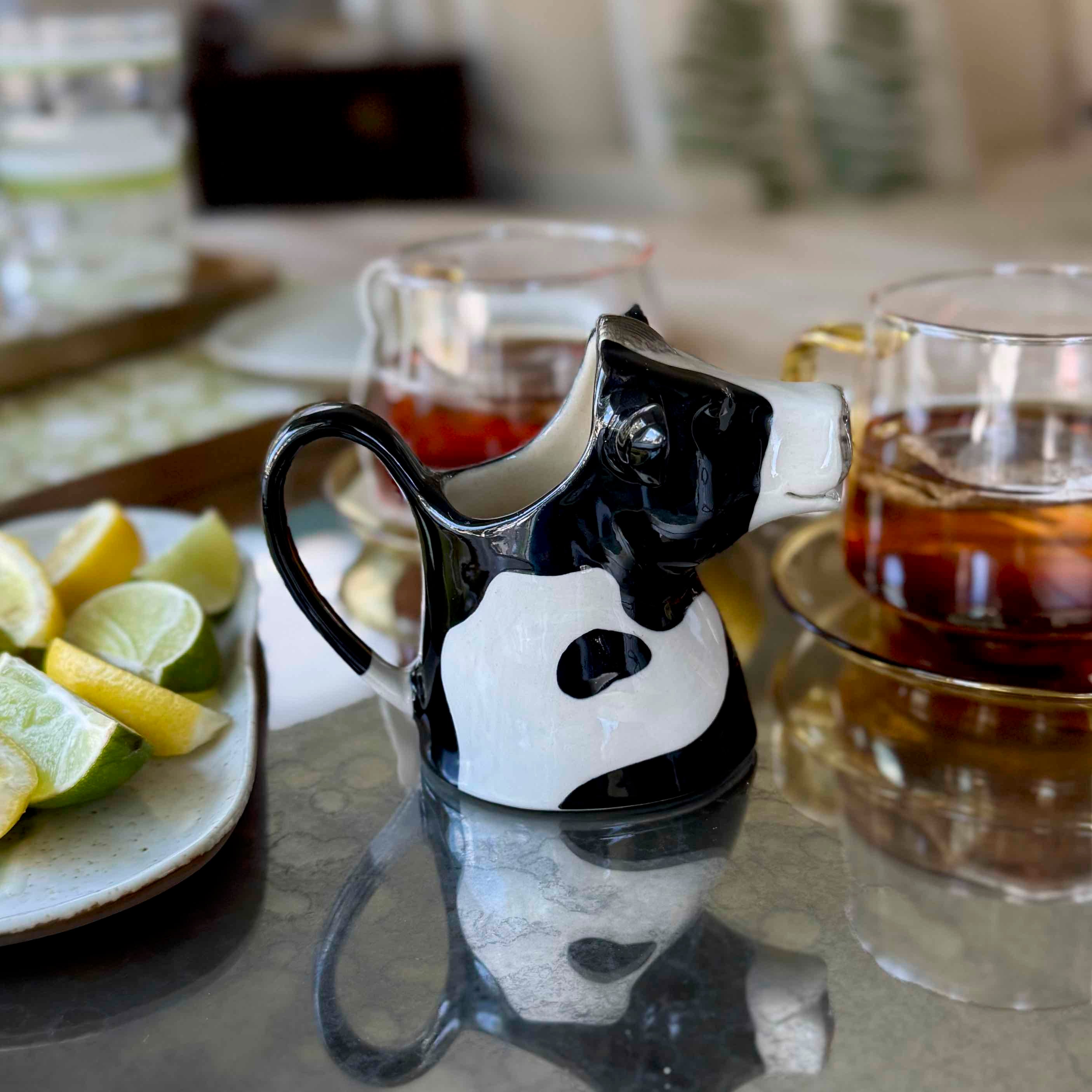 Black and white cow-shaped creamer on a table with tea and fruit.