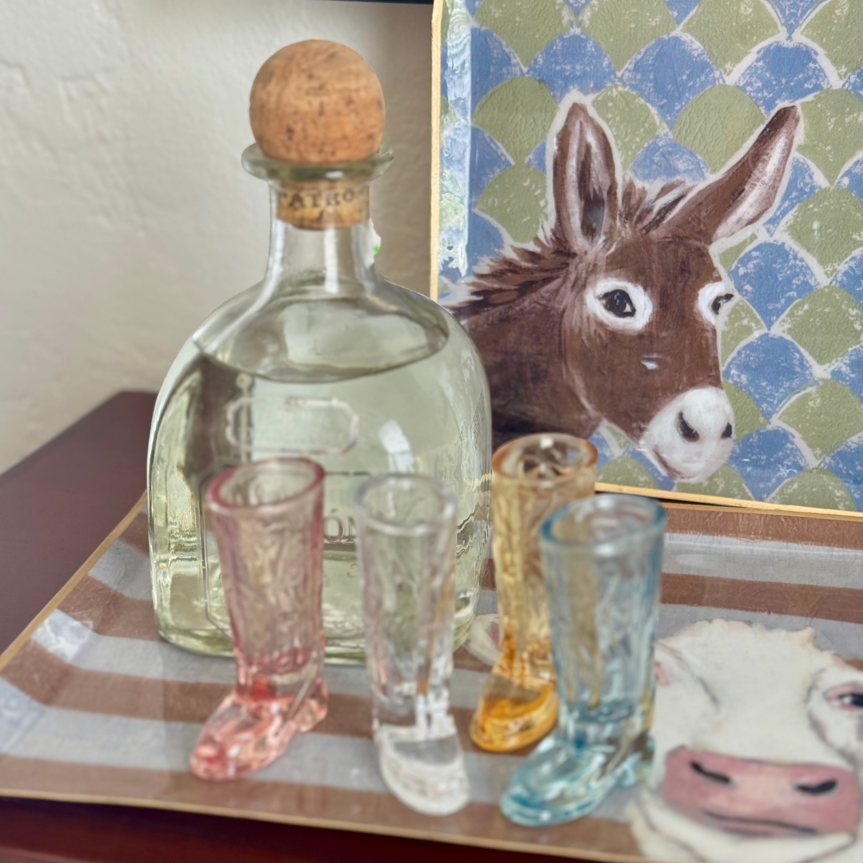 Clear glass bottle with cork stopper on a wooden surface next to colorful shot glasses, with a donkey painting in the background.