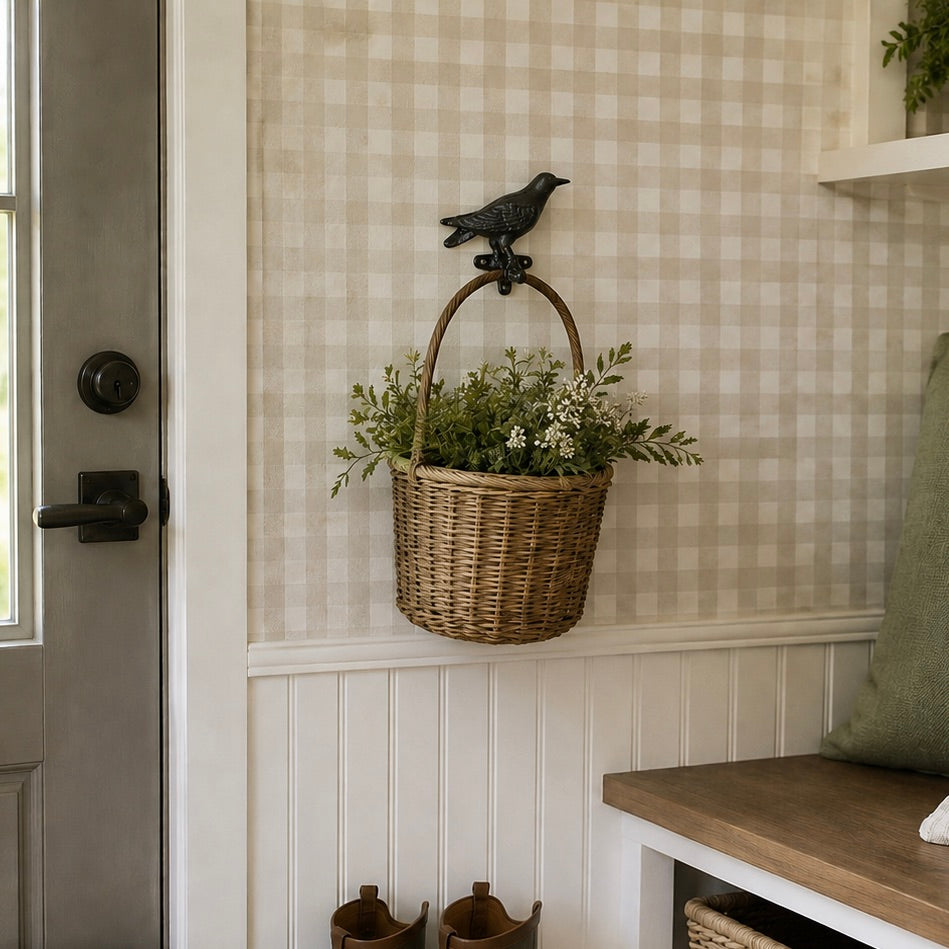 A woven wicker basket with a dark metal bird-shaped hook hanging on a beige-and-white gingham wall in a home entryway. The basket is filled with green plants and sits above white beadboard paneling, with a wooden bench and gray door visible nearby.