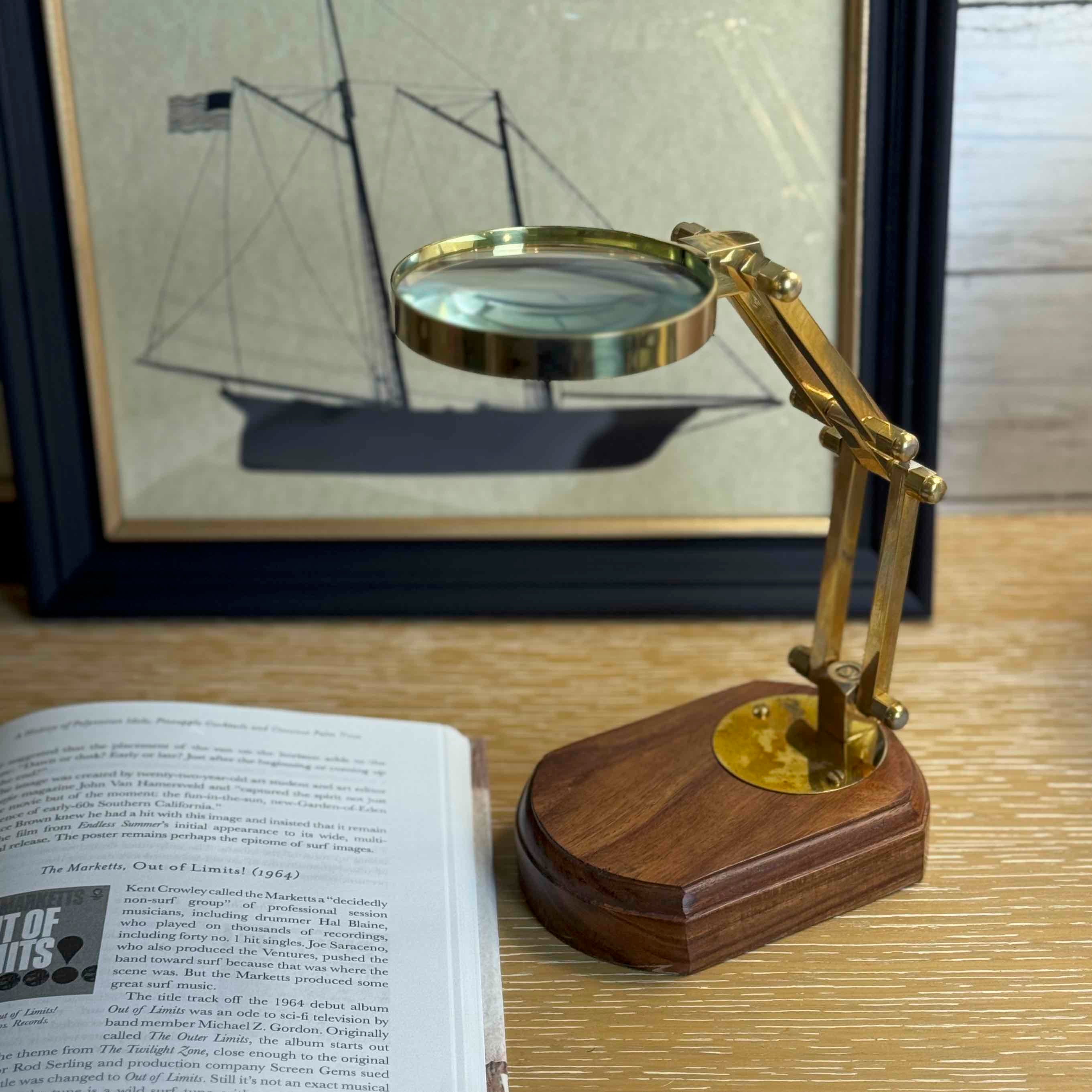 Gold magnifying glass on a wooden base with an open book and framed picture in the background.